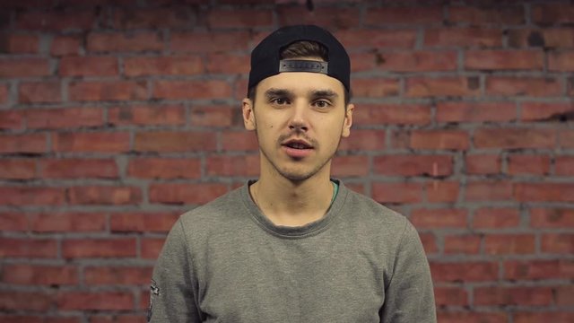 Young man in black cap, grey pullover pronounce words in camera. Brick wall on background. Internet blogger. 