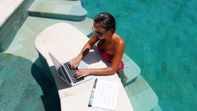 Woman working on her laptop computer sitting at poolside. Beautiful young female model working freelance at swimming pool on laptop.