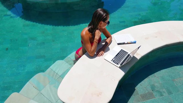 Woman working on her laptop computer sitting at poolside. Beautiful young female model working freelance at swimming pool on laptop.
