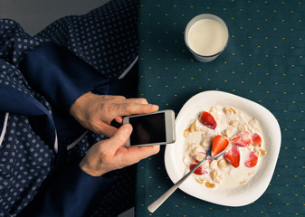 A man has breakfast with milk and strawberries, with phone in hand