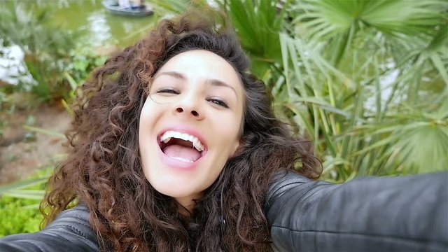 Portrait of happy young woman with beautiful curly hair taking selfie in the park, slow motion