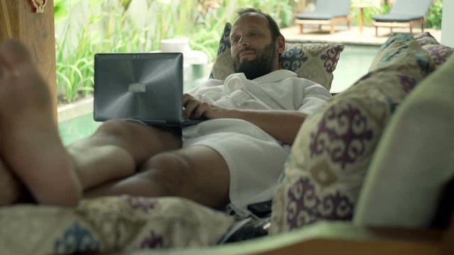 Young man in bathrobe using laptop while lying on sofa in outdoor villa
