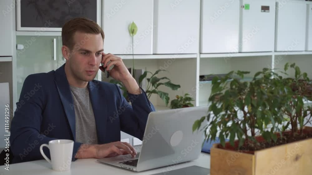 Poster man speaking on mobile phone with client and make notes on the laptop. male in modern office wearing