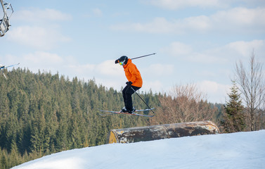Man skier on his skis in flight during a jump over a hurdle from the mountain with firs in the distance on background