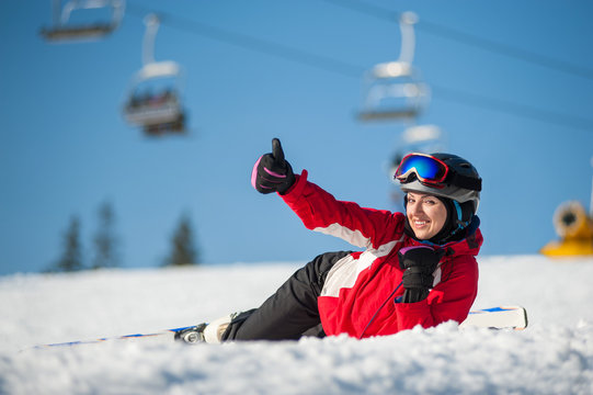 Happy female skier lying with skis on snowy at mountain top, showing thumb up gesture of good class and looking at the camera in sunny day at a winter resort with ski lifts and blue sky in background