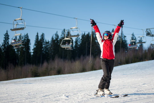 Woman skier wearing helmet, red jacket and ski goggles standing on snowy slope with hands raised up in sunny day with forest and blue sky in background.