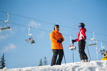 Couple man and woman wearing ski goggles standing with skis on mountain top at a winter resort in sunny day with ski lifts and blue sky in background.