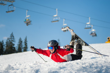 Skier girl in ski goggles lying with raised arms on snowy slope at mountain top in sunny day with...