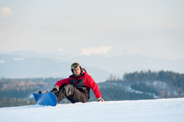 Fototapeta na wymiar Happy snowboarder wearing helmet, red jacket, gloves and pants sitting on snowy slope on top of a mountain, with an astonishing view on hills. Bukovel, Carpathian mountains
