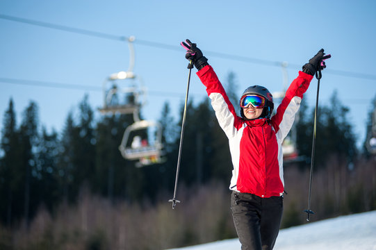 Happy female skier wearing helmet, red jacket and ski goggles standing on snowy slope with hands raised up in sunny day with forest and blue sky in background. Carpathian, Bukovel, Ukraine