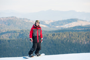 Fototapeta na wymiar Cool snowboarder wearing helmet, red jacket, gloves and pants standing on top of a mountain and looking at the camera on the background of beautiful Carpathian mountains and forests