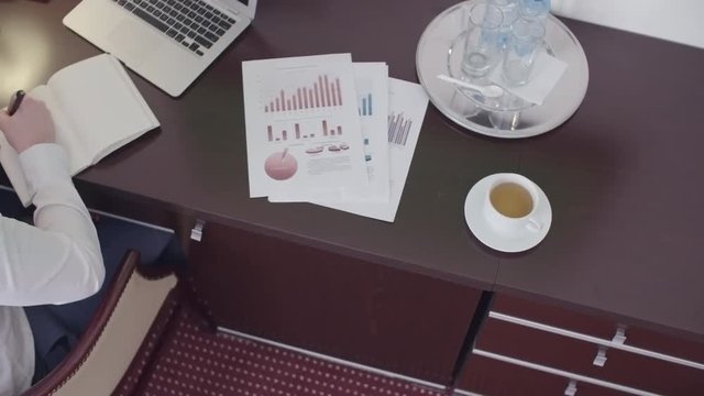 High angle of businessman sitting at the table in hotel room and writing something in his organizer. There is cup of tea, financial documents and laptop on the table