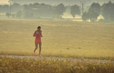 Fit young lady in red summer sportswear running, jogging on a country road while listening to music in earphones, early in the morning. Calories burning. Health care. Willingness concept.