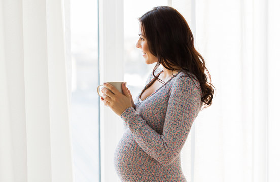 close up of pregnant woman with tea cup at window