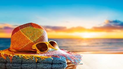 Colorful straw hat,sunglasses and towel, on the wooden table. Summer beach sunrise or sunshine in the background.  