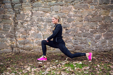Attractive blonde middle aged woman stretching her bodyin front of ancient wall in park