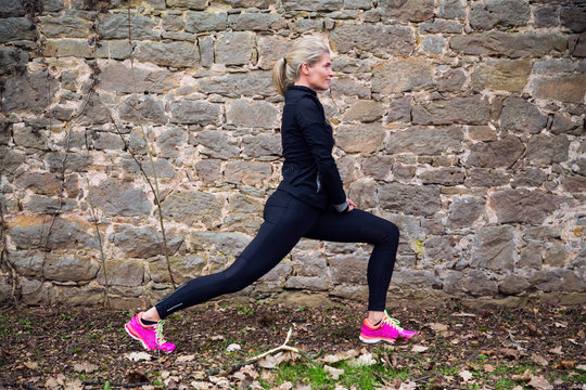 Attractive blonde middle aged woman stretching her bodyin front of ancient wall in park