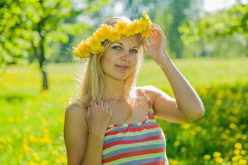 Beautiful, lovely and young woman in the meadow with yellow dandelions and wreath on head in spring sunlight.
