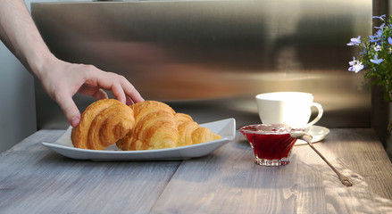 Young man taking croissant for breakfast, Wooden and steel background. Sunny morning.