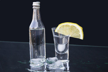 Vodka. Small bottle of vodka with a glass, ice and lemon on the table.  black background, closeup