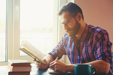 Bearded man writing with pen and reading books at table