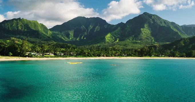 Aerial view flying over hawaiian canoes towards beautiful green mountains and white sandy beach. Hanalei Bay, Kauai