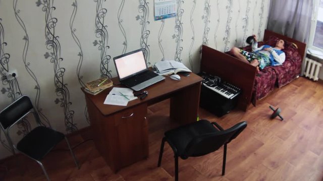 Young boy lay on old bed and throws cap in air in dormitory room. Table with notebook and papers. Student