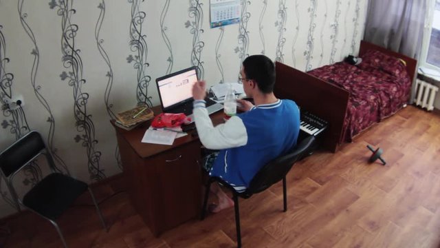 Young boy sitting at notebook in dormitory room and drink milk from bottle, eat cookies. Old bed with red blanket. Synthesizer. Student