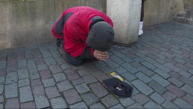 Dolly shot of beggar drifter on streets praying for money a passer by giving coins throwing them in hat laying on old street man kneeling forward does prayer when gets cash stabilized 4k resolution