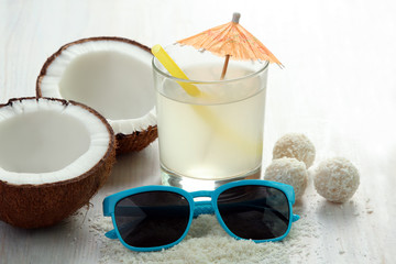 cut the coconut candy coconut near the glass of juice and sunglasses on white wooden background