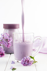 Glass cup with pouring bilberry milkshake near lilac flowers