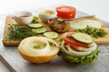 Cooking process of a sandwich burger, inredients on wooden cutting board on wooden table against white background, fresh vegetables, herbs, fried meat, buns, sauces and knife