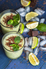 Glasses of kiwi smoothie with mint and chocolate flakes, ice cubes and lime slices on wooden table.