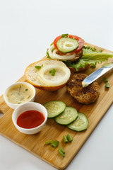 Cooking process of a sandwich burger, ingredients on wooden cutting board on wooden table against white background, fresh vegetables, herbs, fried meat, buns, sauces and knife
