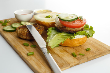 Cooking process of a sandwich burger, ingredients on wooden cutting board on wooden table against white background, fresh vegetables, herbs, fried meat, buns, sauces and knife