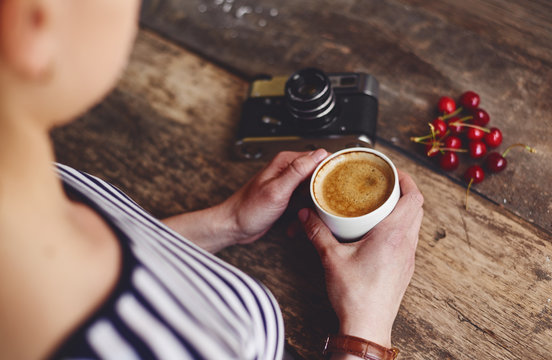 Young woman holding cup of coffee sitting at wooden table with old vintage camera and red cherries. 