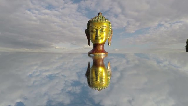 Golden Buddha head sculpture on mirror under cloudy sunny sky, time lapse 4K