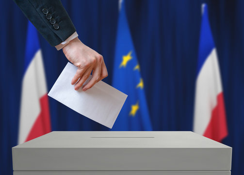 Election in France. Voter holds envelope in hand above vote ballot. French and European Union flags in background.