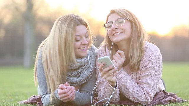 Laughing student girls with earphones lying on grass and singing