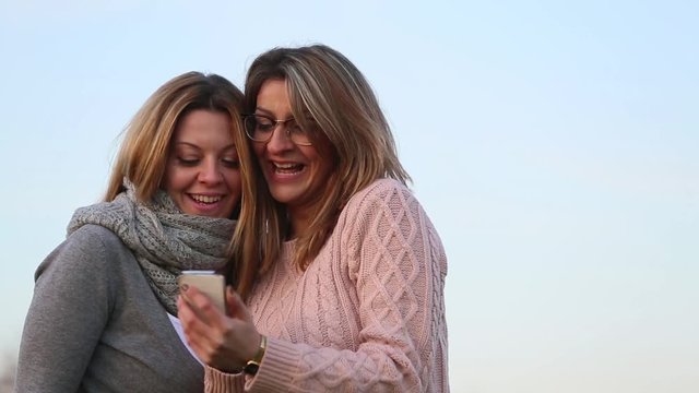 Two simpering girls taking selfie photo and laughing looking at the result. Outdoors