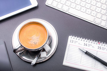 Gefüllte Kaffeetasse umrahmt von Tastatur, Smartphone, Tablet, Kalender und Kugelschreiber