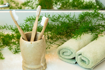 Three bio-degradable, compostable bamboo toothbrushes in marble cup. Rolled green towels in a spa setting. Green plant decor in background. Bathroom white countertop.