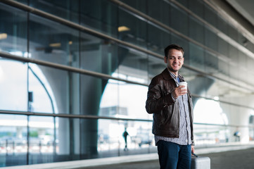 Fototapeta premium Stylish handsome young male traveller with bristle standing outdoors. Man wearing jacket and shirt. Smiling person looking to camera holding cup of coffee