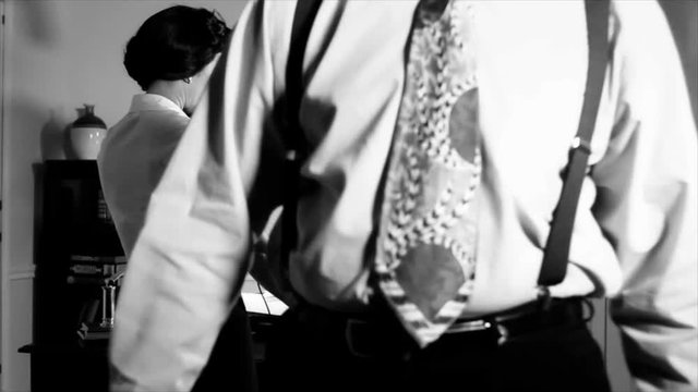 A scene set in the 1940's of a woman making a phone call from a vintage rotary phone while a man leaves the room walking straight towards the camera. Black and White  Film Noir Genre