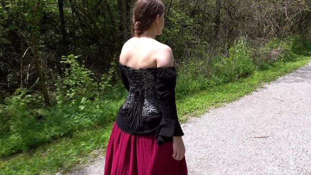 Female dressed up in Renaissance clothing walking through woods on sunny day.