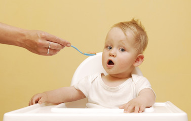 Portrait Of Happy Young Baby Boy In High Chair. The baby boy eating with spoon at home