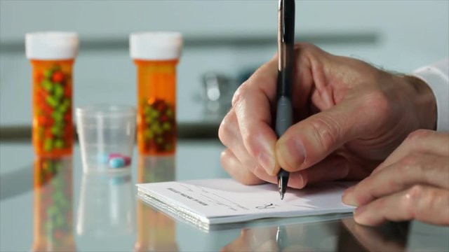 A close up of a male physician's hand writing out a prescription with out of focus pharmaceuticals pill bottles in the background.