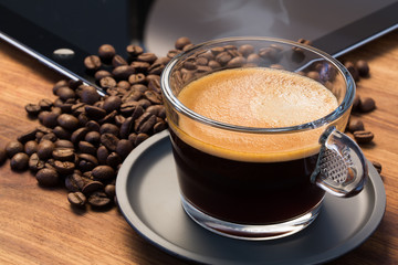 A transparent glass cup of hot steamy espresso on a saucer standing on a wooden table top, surrounded by coffee beans and an ipad in the background