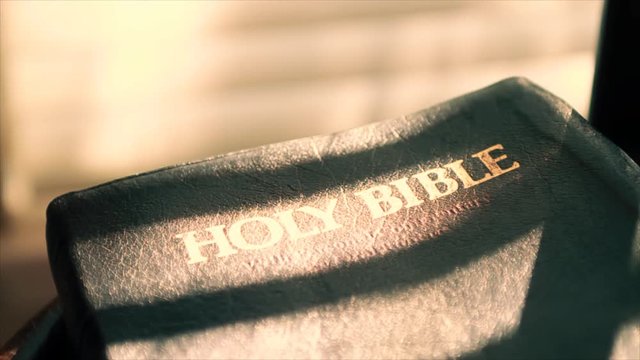 A woman's hand reaches down and picks up from a chair a Bible that is bathed in the sunbeams filtering through window shades.