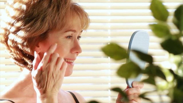 A mature woman looking in a mirror smiles briefly then becomes dissatisfied with her reflection and begins to examine her aging skin.